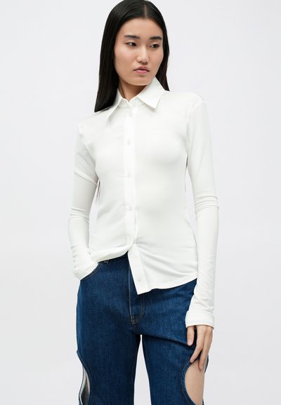 Camisa blanca de manga larga con botones y cuello, confeccionada en una tela suave. Presenta un diseño entallado y se lleva con detalles de jeans azules.