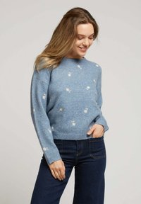 Pull bleu clair en tissu doux avec un col rond, présentant une broderie florale blanche. Coupe ample et poignets côtelés.