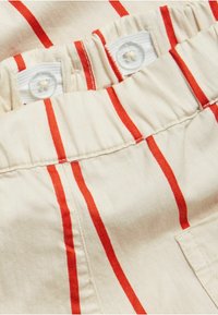 Beige kangas yenye mistari ya wima ya rangi nyekundu, ikiwa na waistband inayoweza kunyumbulika iliyo na vifungo viwili vyeupe. Muonekano unaonekana kuwa laini na mwepesi.