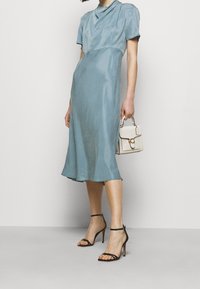 Kvinna i en himmelsblå satin klänning som når till mitten av låren, svarta högklackade sandaler med remmar, och håller en liten vit handväska med gulddetaljer.