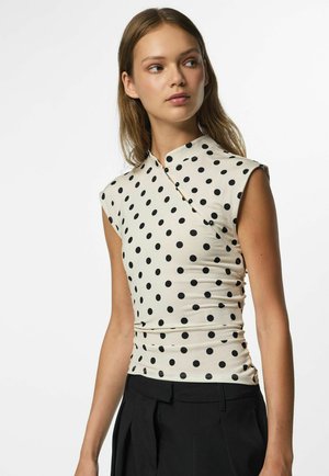 Jeune femme portant un haut blanc sans manches à pois noirs et un pantalon noir, regardant sur le côté sur un fond uni.