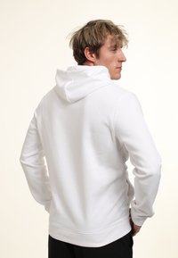 Giovane uomo con capelli biondi che indossa una felpa bianca semplice, visto di spalle con le mani sui fianchi su uno sfondo neutro.
