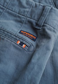 Pantaloni di cotone azzurri chiari con una tasca sul retro decorata con una striscia arancione e blu scuro e un'etichetta con il logo "SUPERDRY" in rosso e blu.
