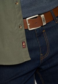 Camicia verde oliva con bottoni a scatto e un'etichetta logo bordeaux, abbinata a jeans blu scuro con cuciture gialle a contrasto e una cintura in pelle marrone.