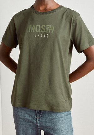 Man in een olijfgroene t-shirt met MOSH-logo en blauwe jeans, staand met zijn handen achter zijn rug tegen een effen achtergrond.