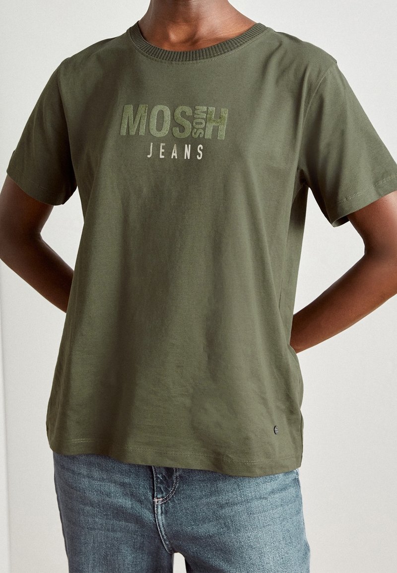 Un homme portant un t-shirt de marque MOSH de couleur vert olive et un jean bleu, debout avec les mains derrière le dos devant un fond uni.