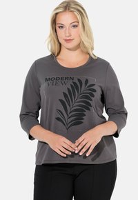 Vaaleahiuksinen nainen, joka käyttää harmaata 3/4-hihaista paitaa mustalla lehtikuvioinnilla ja tekstillä "MODERN VIEW", seisoo kädet yhteen sidottuina.