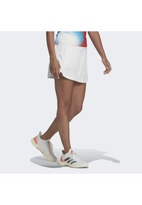 Witte sportrok met een gebogen zoom, gecombineerd met lichtgrijze sneakers met zwarte strepen en koraalaccenten. Gladde stof, actieve pasvorm.