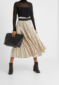 Top lungo trasparente nero a maniche lunghe, gonna plissettata beige a scacchi, stivali neri alla caviglia e una grande borsa a tracolla nera, evidenziando linee e texture pulite.