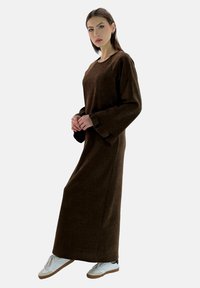 Vestido largo de manga larga en marrón, hecho de una tela suave con mangas acampanadas, combinado con zapatillas blancas con suelas marrones. Textura suave, hasta el suelo.