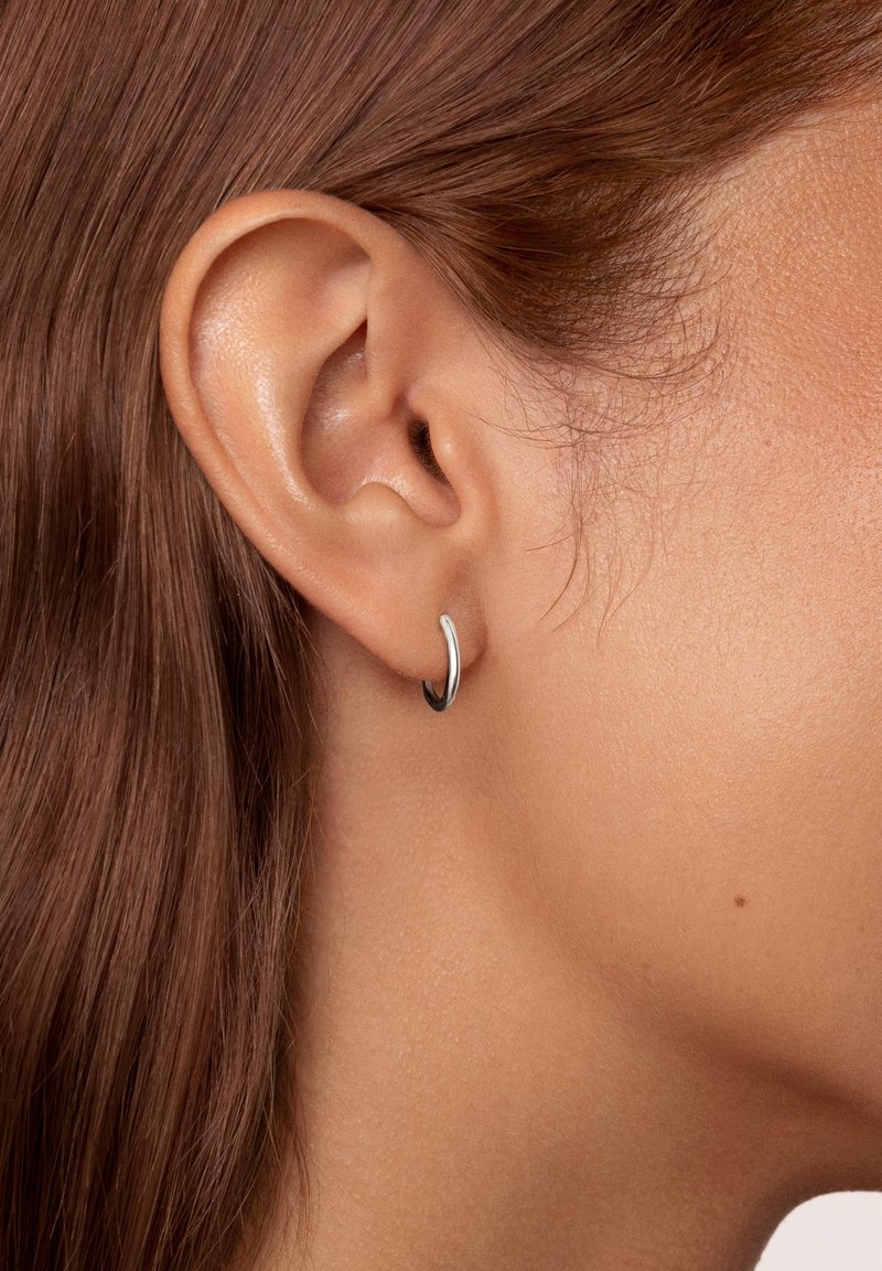 Orecchino a cerchio in argento con un design liscio e arrotondato. Superficie riflettente, dimensioni medie, si adatta perfettamente al lobo dell'orecchio su una pelle chiara.