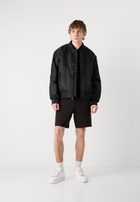 Giacca bomber nera in materiale lucido, con colletto rialzato e chiusura con zip. Abbinata a pantaloni corti neri e sneakers bianche.