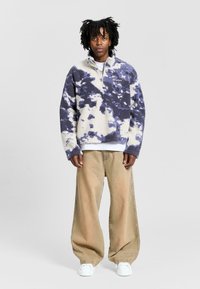 Fleece pullover with purple and cream abstract pattern, half-zip neck, and long sleeves. Worn with beige wide-leg pants and white sneakers.