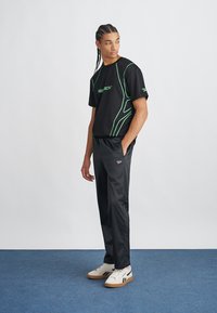 Top preto de mangas curtas com detalhes verdes, combinado com calças desportivas pretas e ténis brancos. Modelo em pé contra um fundo claro.