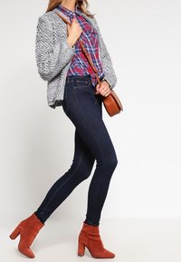 Vrouw in een grijze gestructureerde cardigan, rode geruite overhemd, donkere skinny jeans, rode enkellaarsjes en met een bruine schoudertas.