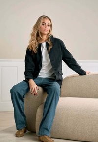 Sort jakke over en hvid skjorte, parret med blå jeans og beige sko. Uformel setting med en lysfarvet, tekstureret sofa.