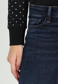 Svart topp med vit prickmönster vid ärmarna, tillsammans med mörkblå denimjeans. Närbild på handen vid höftområdet.