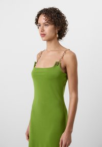 Robe en matériau extensible vert avec un décolleté carré, présentant des accents en chaîne dorée sur les bretelles. Texture lisse et design moulant.