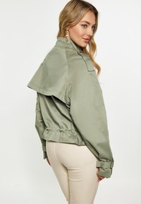 Jachetă scurtă de culoare verde măslin cu guler larg, manșete cu curea și talie elastică. Textură netedă, cu o finisare mată și fără închizători vizibile.