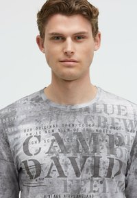 Graues Langarmshirt mit strukturiertem, verblasstem Design und schwarzem Text, darunter "CAMP DAVID", in unterschiedlichen Schriftarten und -größen.
