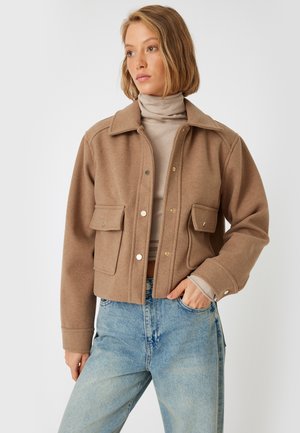 Giacca corta marrone in morbido misto lana, con chiusura a bottoni, colletto a punta e due tasche frontali. Indossata sopra un dolcevita beige chiaro.