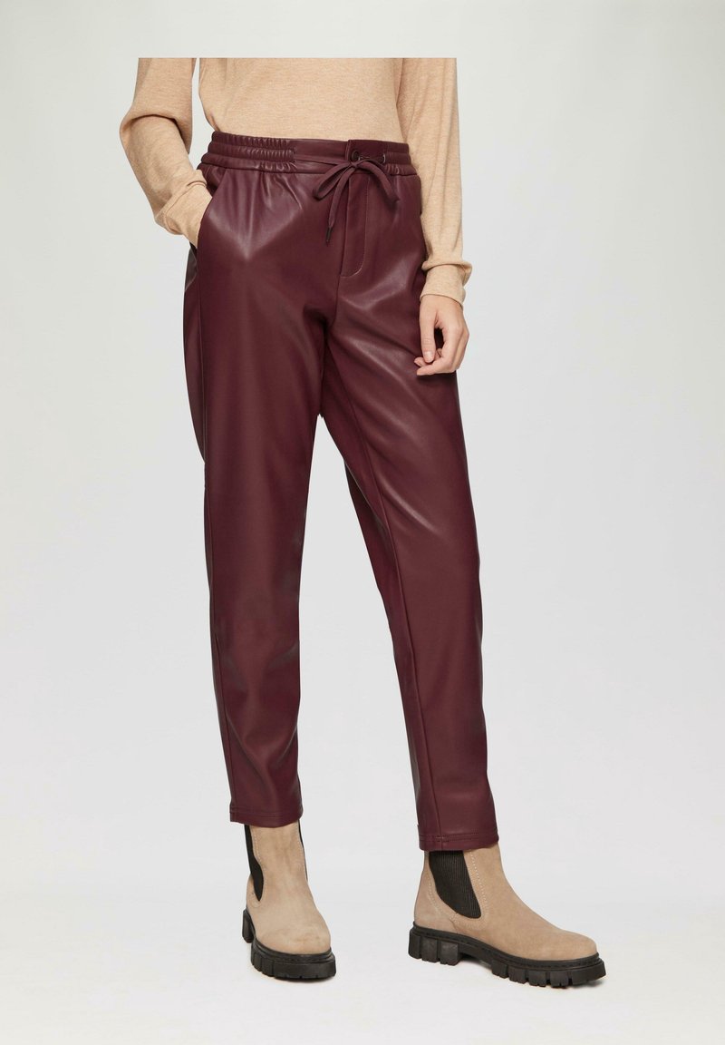 Žena má na sobě burgundské jogger kalhoty z umělé kůže, béžový dlouhý rukáv a béžové kotníkové boty s černou elastickou stranou.