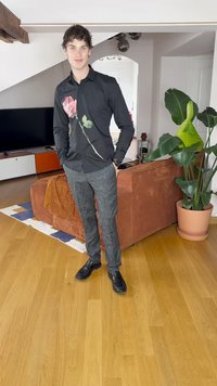 Sort skjorte med knapper og rose-design på venstre side, parret med mønstrede mørkegrå bukser og sorte sko. Baggrund med trægulv.