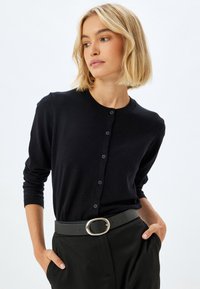 Cardigan nero con scollatura rotonda, maniche lunghe e chiusura con bottoni. Indossato sopra pantaloni neri a vita alta, abbinato a una cintura con fibbia argentata.