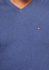 Gros plan sur un pull bleu à col en V avec un petit logo rectangulaire aux couleurs blanc, rouge et noir sur la poitrine.