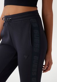 Zwarte sportbroek met een koord in de taille, een zijzak en een glanzende "GUESS" logo-streep die langs de zijkant loopt. Gladde textuur en aangesloten ontwerp.