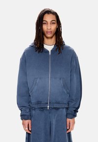 Νεαρός με μακριά dreadlocks, που φοράει μπλε φούτερ με φερμουάρ και παντελόνι που ταιριάζει, στέκεται μπροστά σε απλό φόντο.