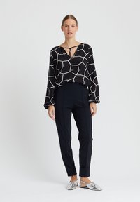 Zwarte blouse met een wit geometrisch patroon, met een gestrikte halslijn, gecombineerd met donkere broeken en zilveren instappers.
