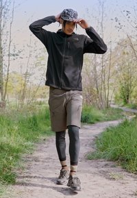 Zwarte zip-up jas, olijfgroene shorts, zwarte leggings, patroon pet, en zwarte schoenen. Man die over een pad wandelt omringd door groen.