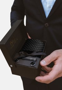 Schwarze Geschenkbox mit Logo, gefüllt mit einer gepunkteten Krawatte, einem silbernen Stift und Manschettenknöpfen. Die Box ist mit einem braunen Band gebunden. Glatte Oberfläche.
