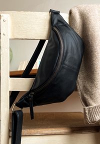 Sac bandoulière en cuir noir suspendu à une chaise en bois blanc usée avec un pull en tricot beige drapé sur le dossier.