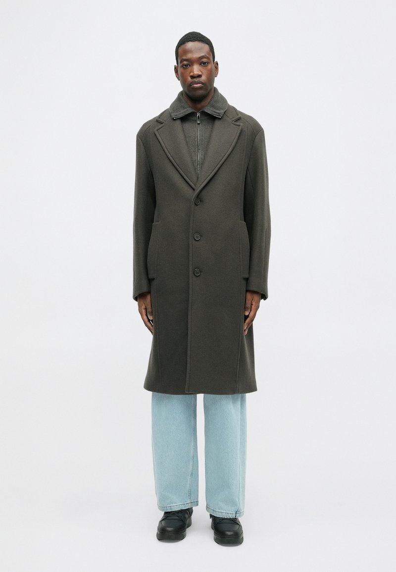 Tamsiai žalia ilga striukė su dideliais atlapais, dviem priekinėmis kišenėmis ir trimis saga. Dėvima ant tekstūruoto megztinio ir šviesiai mėlynų džinsų. Juodi batai.