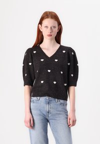 Maglione nero lavorato a maglia con scollo a V e maniche corte a palloncino, con motivo a cuori bianchi. Abbinato a jeans in denim azzurro chiaro.
