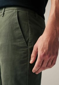 Dark green trousers made from smooth fabric, featuring a flat front, side pockets, and a close-up of a hand resting on the pant leg.