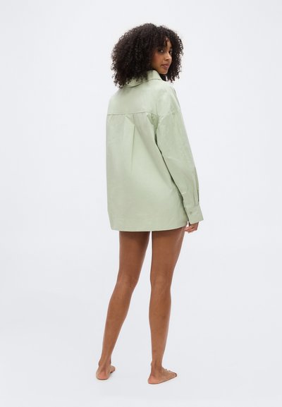 Γυναίκα με σγουρά μαλλιά που φοράει ένα φαρδύ ανοιχτό πράσινο πουκάμισο, όρθια ξυπόλητη, γυρισμένη ελαφρώς στο πλάι μπροστά σε απλή φόντο.