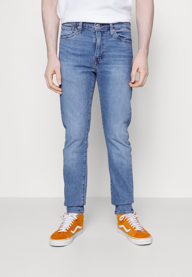 Jeans de denim azul claro con un corte clásico, cinco bolsillos y un ligero desvanecimiento; combinados con zapatillas de lona naranjas y blancas.