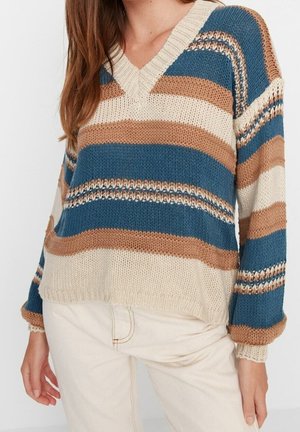 Frau trägt einen gestrickten V-Ausschnitt-Pullover mit breiten horizontalen Streifen in Blau, Beige und Braun, kombiniert mit hellbeigen Hosen.