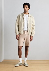 Φωτεινό μπεζ βomber μπουφάν, λευκό μπλουζάκι και μπεζ σορτς. Πράσινα και γκρι αθλητικά παπούτσια με λευκές κάλτσες. Απλός σχεδιασμός με ελαστικές μανσέτες.