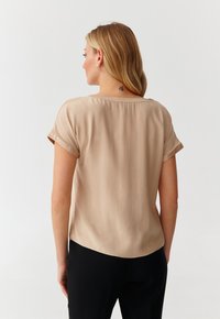 Beige short-sleeve blouse with a round neckline and rolled cuffs, featuring a smooth texture and slightly curved hem. Back view shown.