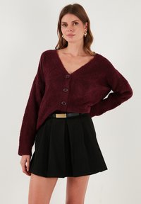 Burgundi színű bolyhos kardigán mély V-nyakkivágással, három nagy gombbal és hosszú ujjal, fekete redős szoknyával és vékony övvel párosítva.