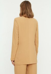 Blazer de manga larga beige con una tela texturizada, que presenta un corte recto y un detalle mínimo, mostrado desde la parte trasera.