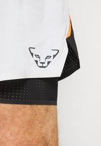 Shorts de sport blancs avec des détails en maille noire, arborant un logo de lion noir bien en vue sur le côté, et une touche d'orange sur l'ourlet.