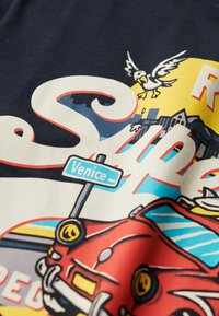 Camiseta marina con un estampado gráfico de un coche rojo, una gaviota y un cartel de calle de Venecia. Algodón suave con una textura lisa. Colores brillantes.
