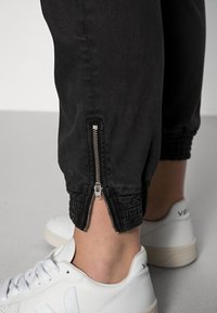 Černé zúžené kalhoty se zipovými lemy a nařasenými manžetami, vyrobené z měkkého materiálu. Spárovány s bílými teniskami, které mají hladkou texturu.