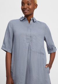Hellblaues Leinenhemdkleid mit Kragen, kurzen aufgerollten Ärmeln, einer Fronttasche und Knopfleiste. Sanfte Textur mit lässiger Passform.
