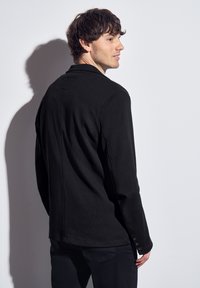 Svart långärmad skjorta i texturerat tyg, med knappdetalj vid muddarna och rak skärning. Bakifrån visas en ren silhuett.